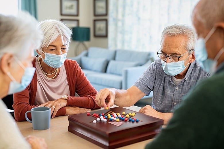 Group of seniors men and women wearing protective face mask playing chinese checkers at nursing home during pandemic lockdown. Old man wearing eyeglasses and surgical face mask for safety against covid-19 playing with a group of friends. Senior people enjoy a board game at care facility centre.
