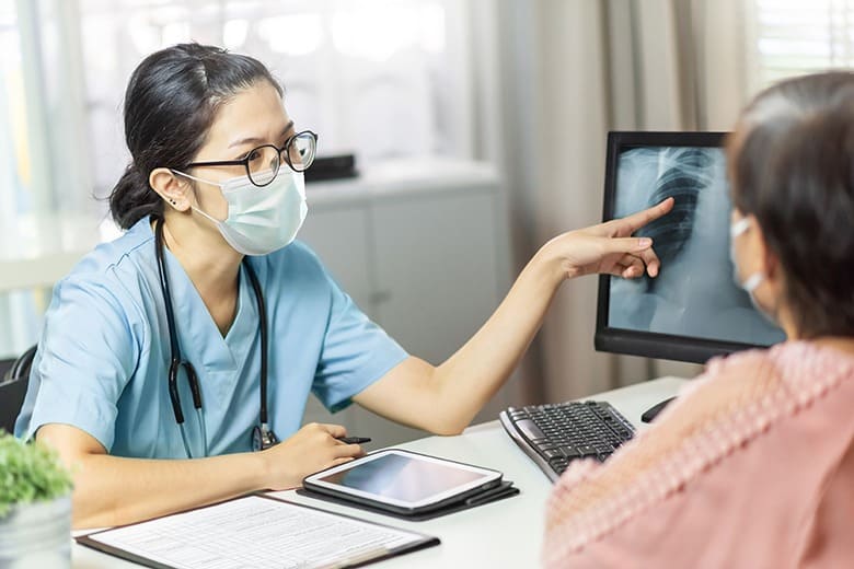 Asian female doctor in medical mask examining and pointing to radiological chest X-ray film on monitor computer while talking with Elderly woman patient at hospital room. Lung radiography