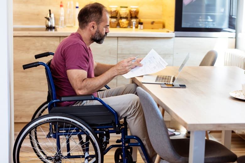 diabled-man-in-wheelchair-reading-paperwork-sitting-in-front-of-laptop