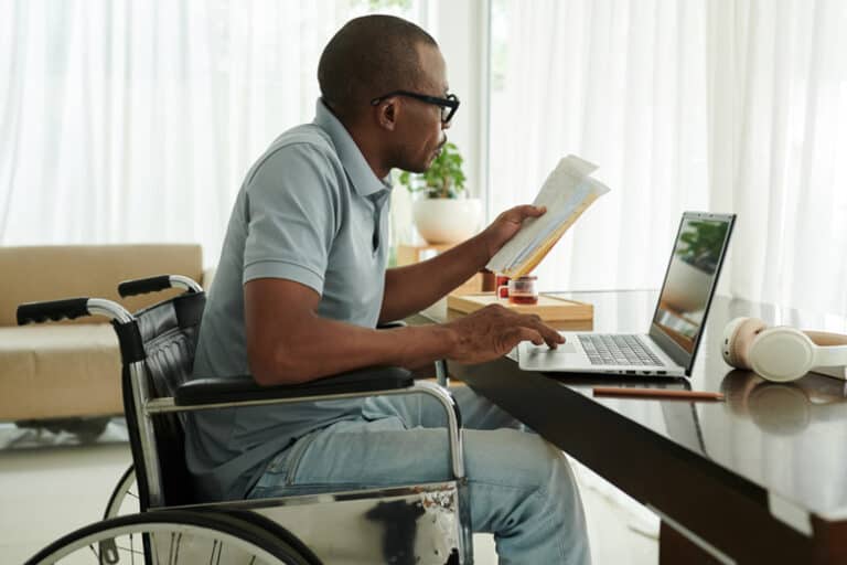 man-wearing-glasses-in-wheelchair-reading-paperwork-on-laptop